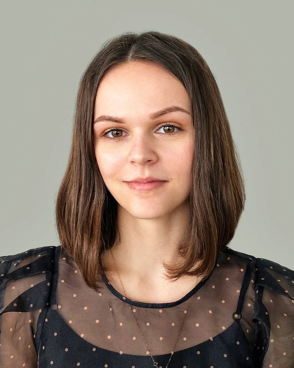 A young woman with shoulder-length brown hair, wearing a black sheer top with pink polka dots, looking at the camera with a slight smile.