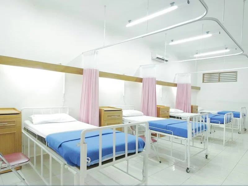 Hospital ward with four beds, each separated by pink privacy curtains, with wooden bedside tables, white walls, and ceiling lights.