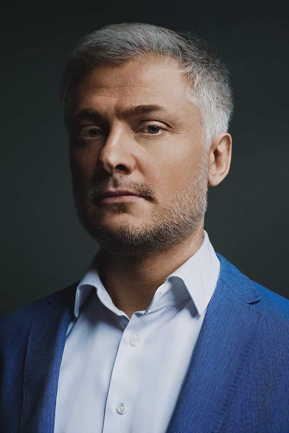 Portrait of a mature man with gray hair and beard, wearing a white dress shirt and blue suit jacket, looking confident against a dark background.