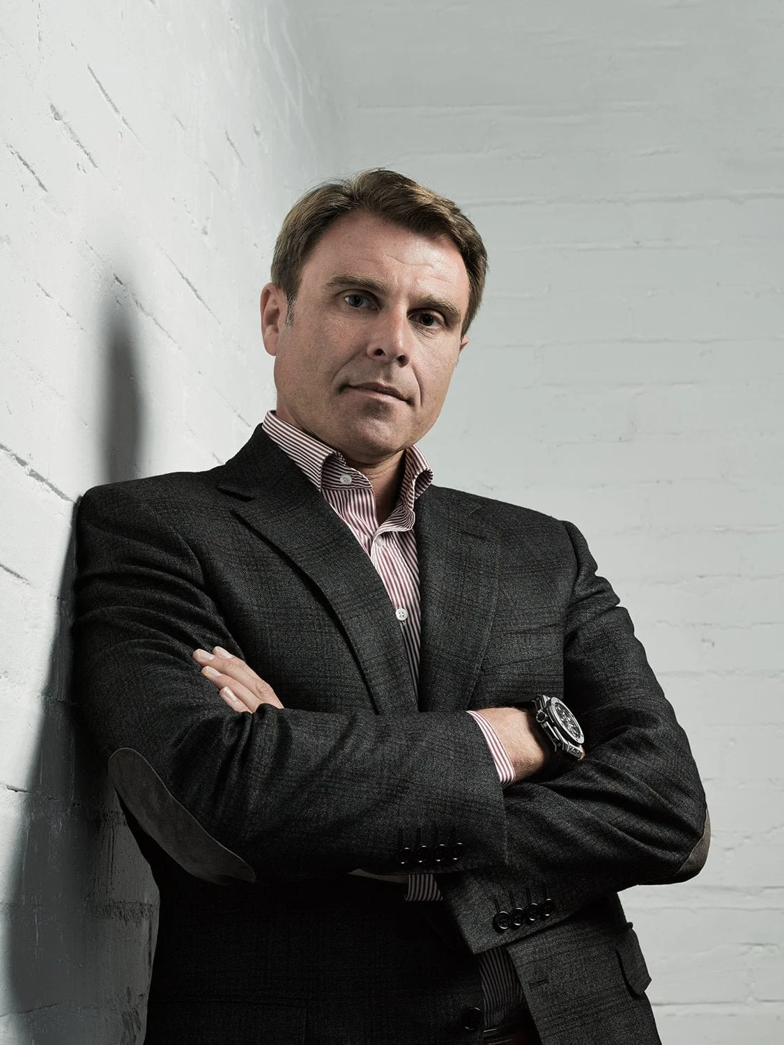 A man in a dark suit with a watch, arms crossed, standing against a white brick wall.