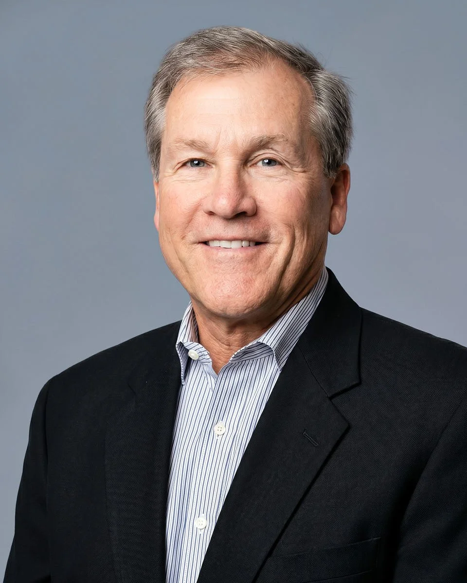 A professional headshot of a smiling middle-aged man with gray hair wearing a dark suit jacket and a light blue striped shirt against a gray background.