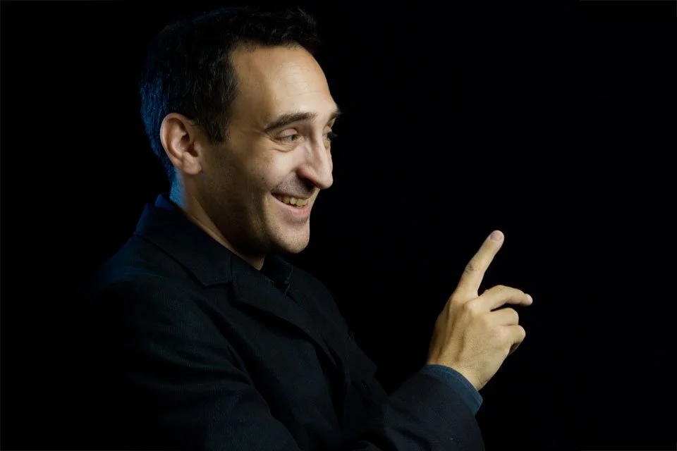 A man smiling and pointing upward with his index finger against a black background.