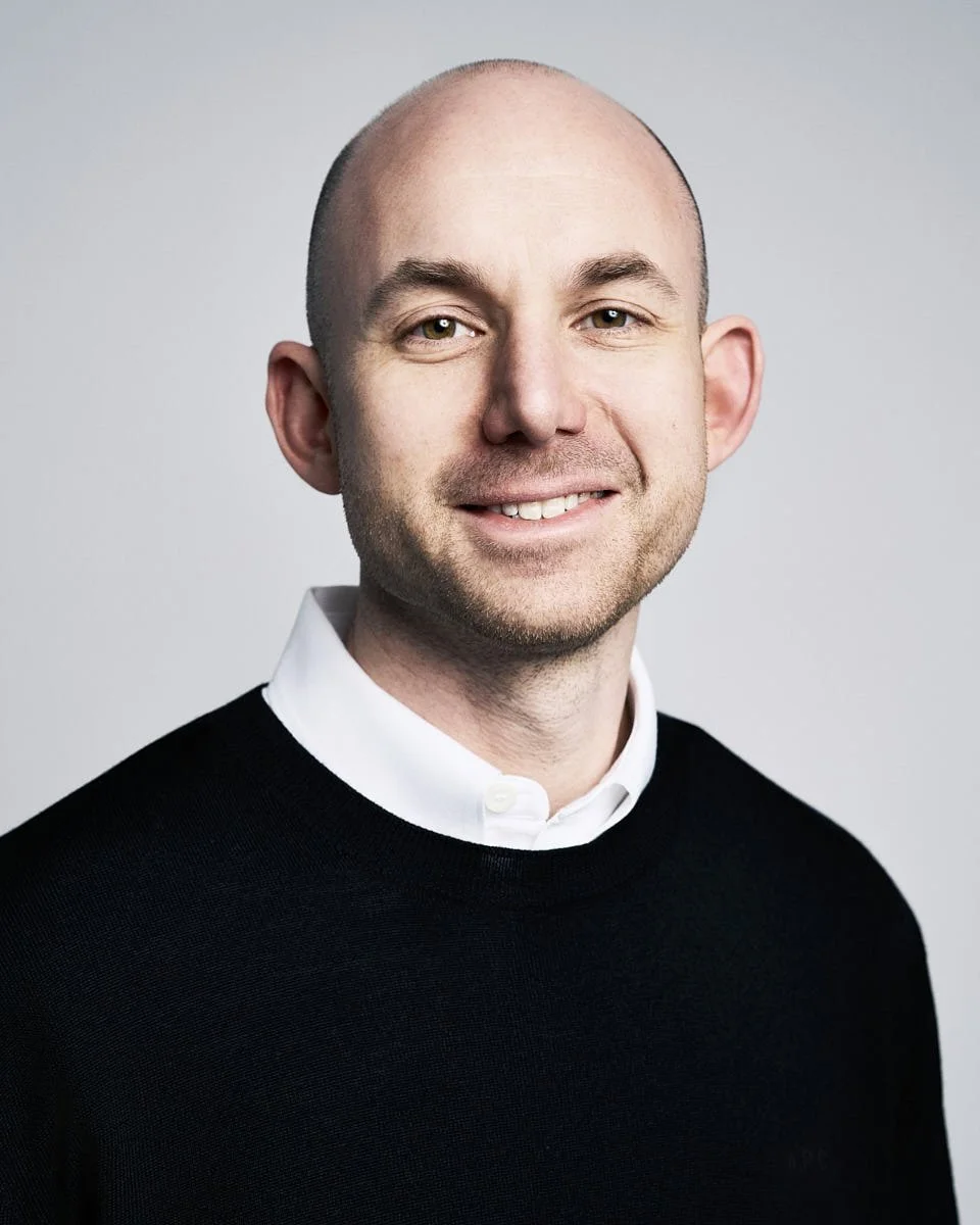 Headshot of a bald man in a white collared shirt and black sweater, smiling against a light gray background by Match Production.