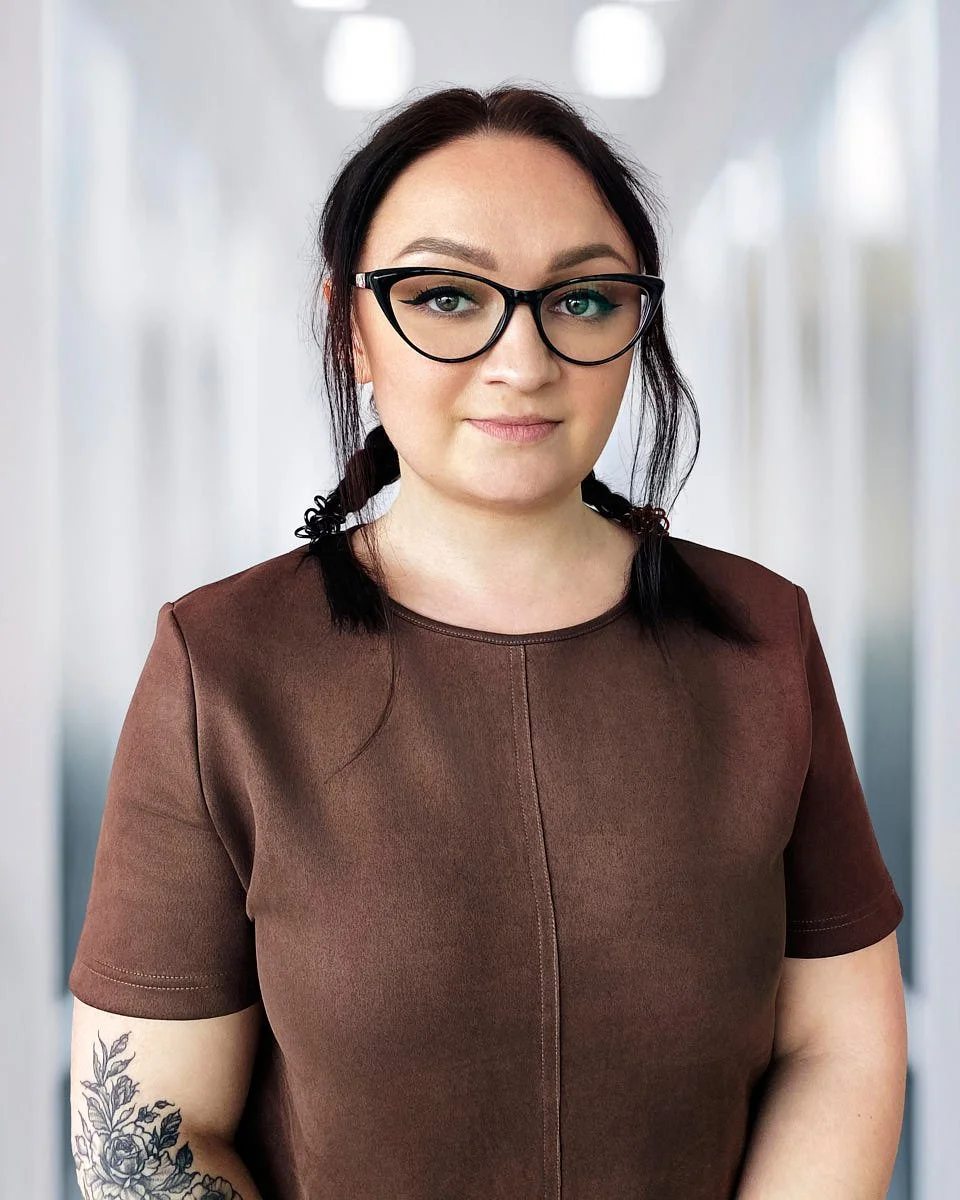 A woman with dark hair in braids, wearing glasses and a brown shirt, standing in a hallway.