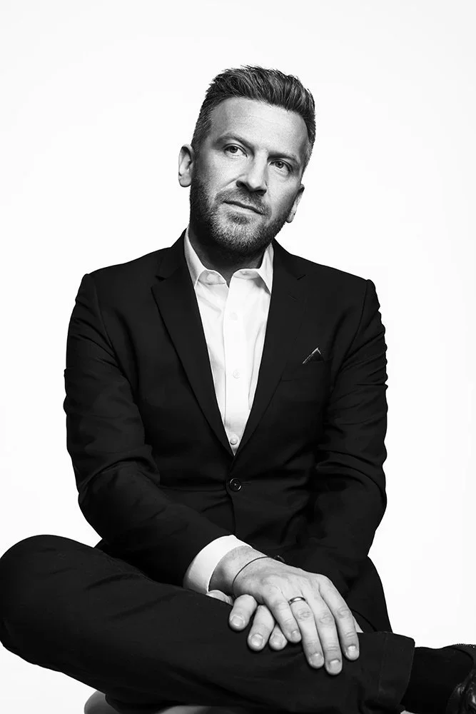 Black and white portrait of a man sitting with legs crossed, wearing a suit and white shirt, with short styled hair and a beard.