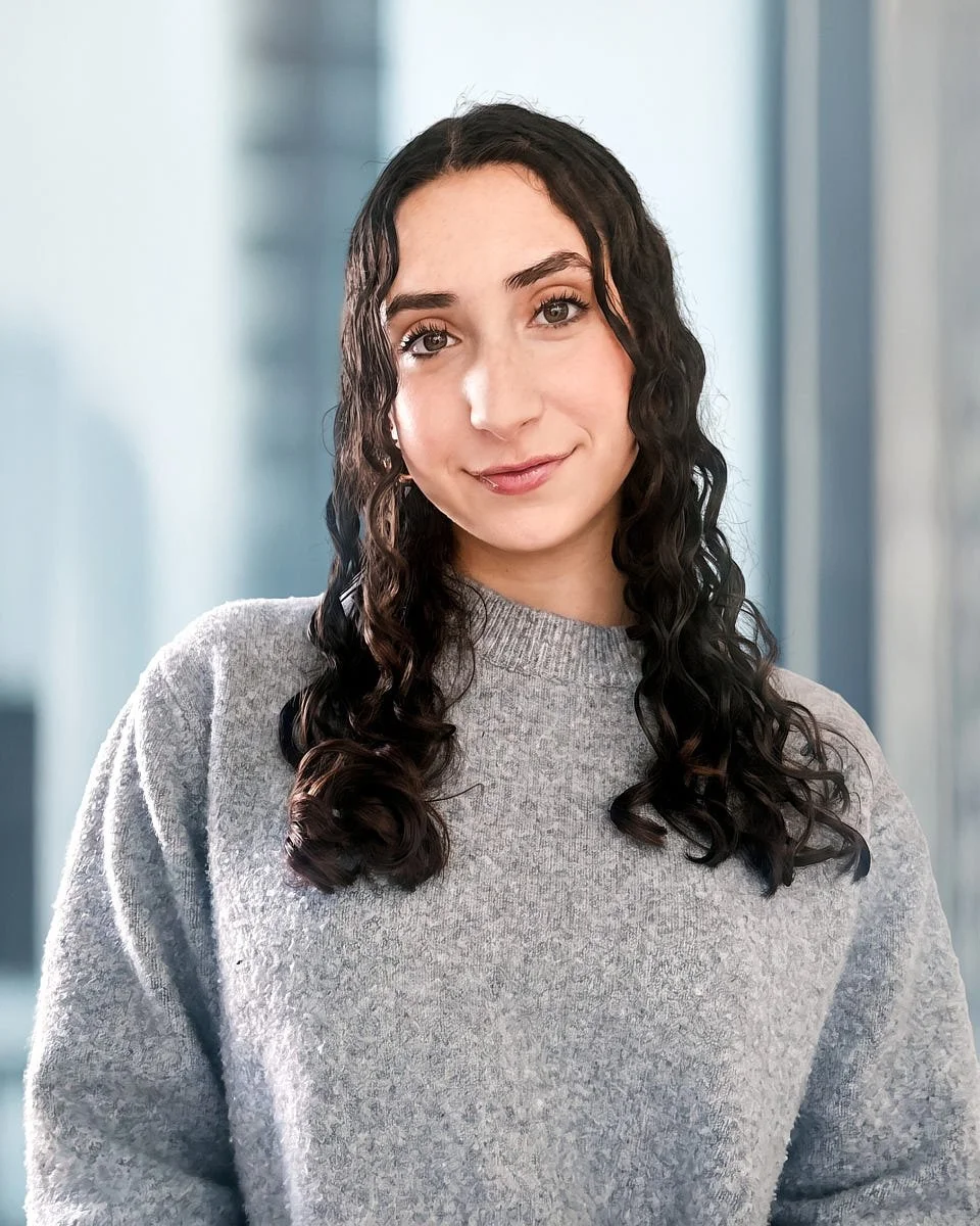 A woman with long, curly dark hair, wearing a light gray sweater, smiling softly indoors with a blurred modern office background.