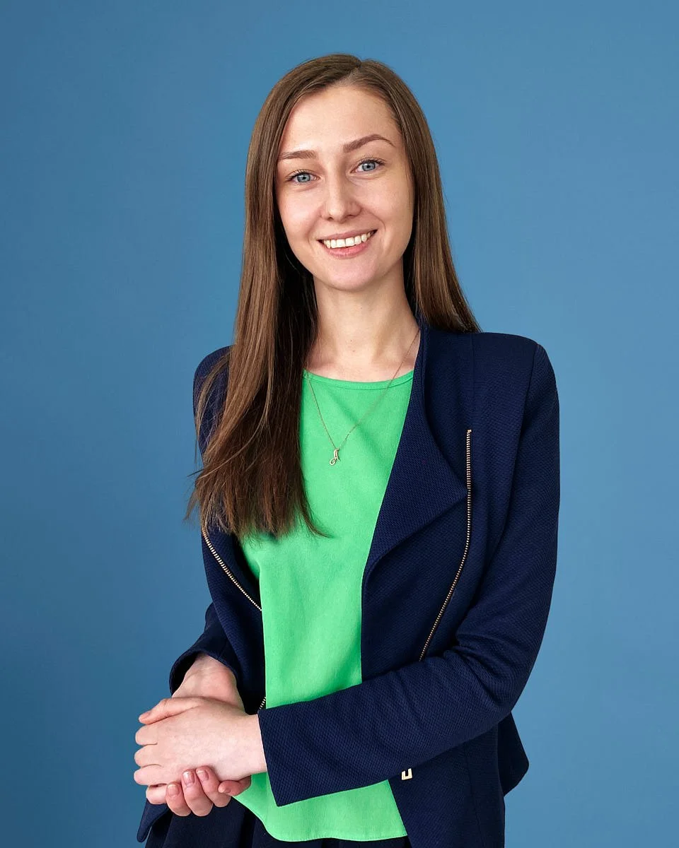 A young woman with long brown hair, blue eyes, and fair skin, smiling while standing against a blue background. She is wearing a green top, a dark blue jacket, and a silver necklace with a small pendant.