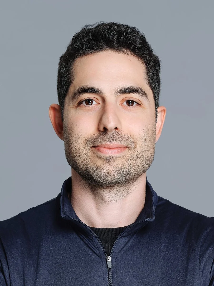 Headshot of a man with dark hair and light skin, wearing a navy blue zip-up jacket, smiling softly against a gray background by Match Production NYC.