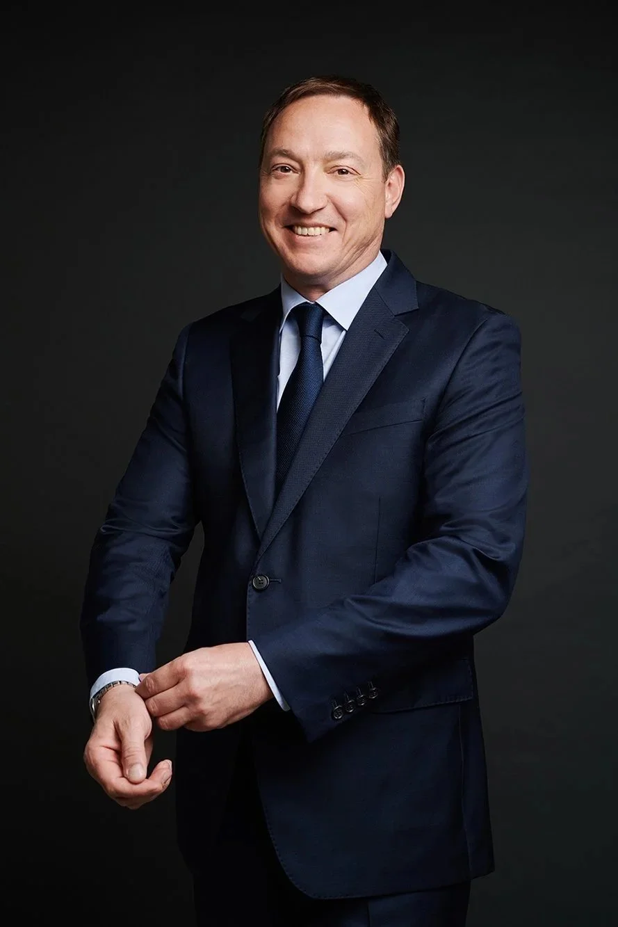 A smiling man dressed in a navy blue suit, white shirt, and navy tie, standing against a dark background.