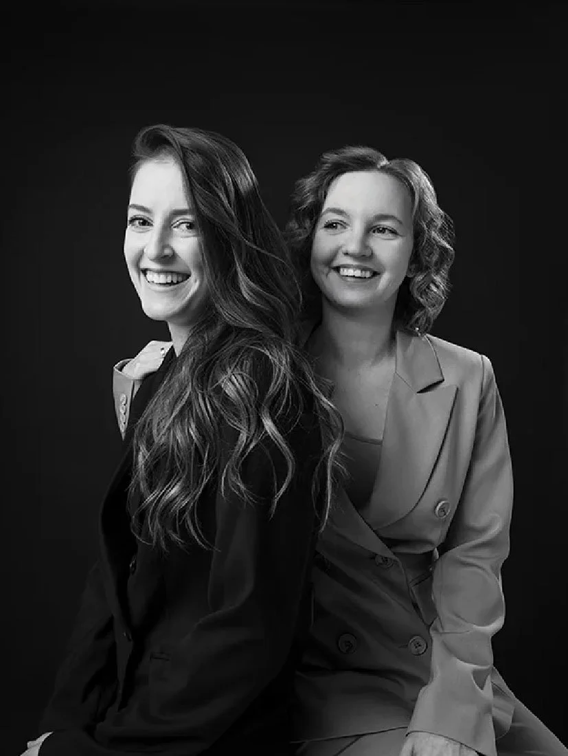 Black and white portrait of two women smiling, one with long wavy hair and the other with short curly hair, sitting against a dark background.