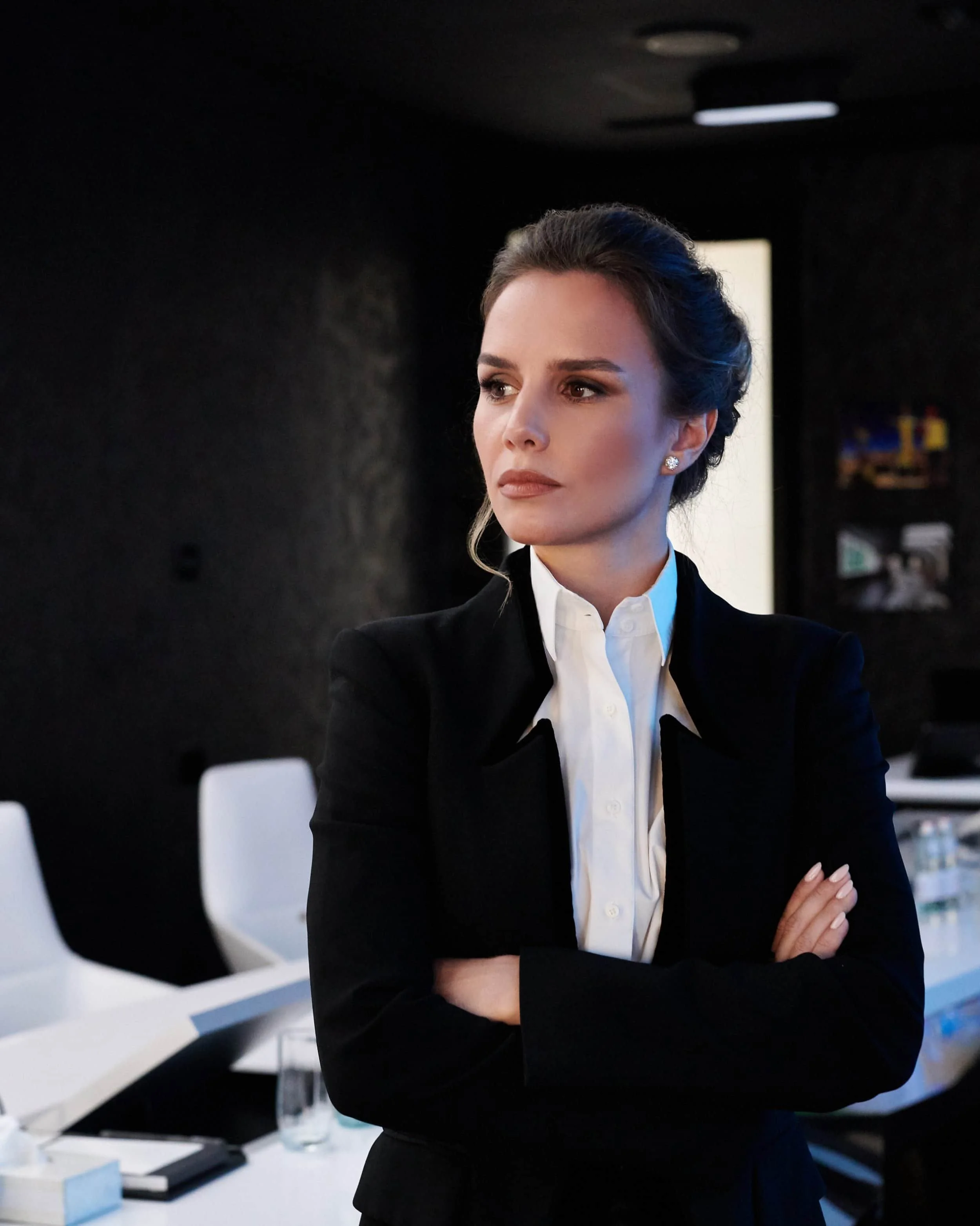 A woman with brown hair in an updo, wearing a black blazer and white shirt, standing with crossed arms in a dark room, looking to the side.