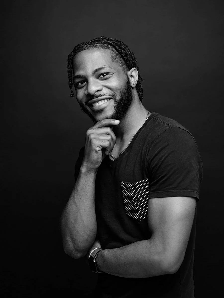 A black and white portrait of a smiling man with dreadlocks and a beard, wearing a dark t-shirt with a polka dot pocket, posing with his hand on his chin against a dark background.