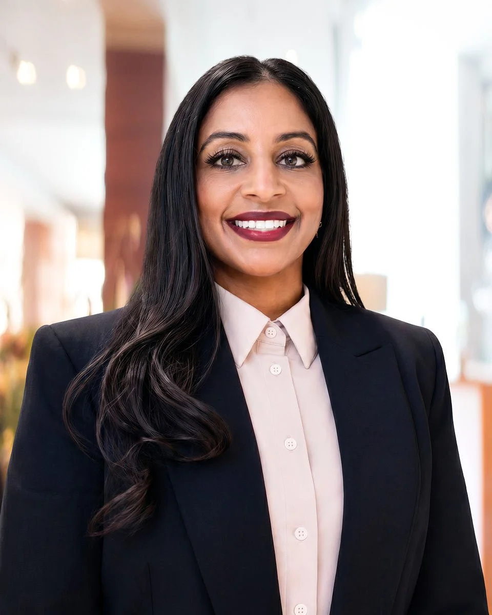 Professional woman with long dark hair smiling in an office setting, wearing a black blazer and light-colored shirt.