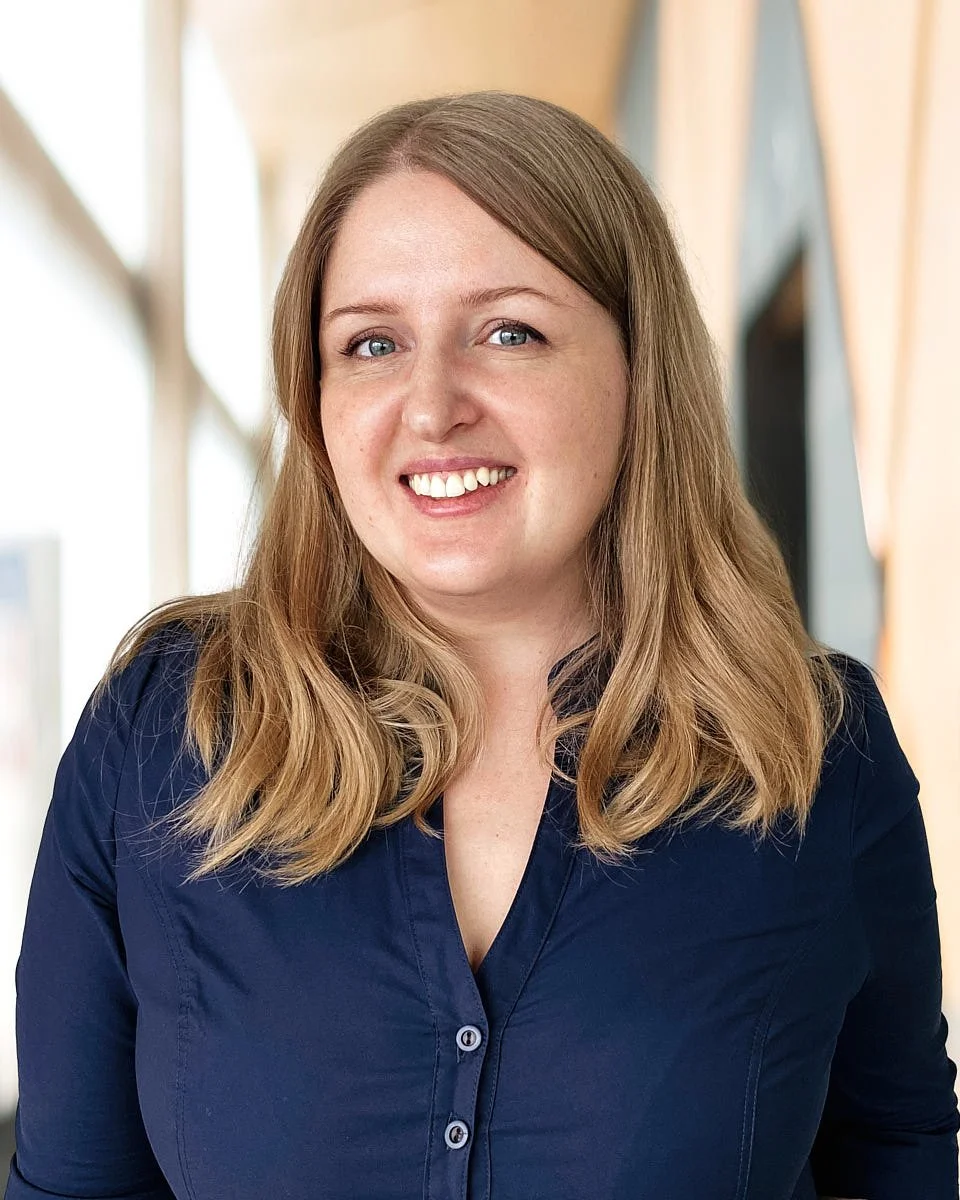 A woman with shoulder-length light brown hair, wearing a navy blue blouse, smiling in a well-lit indoor space with large windows in the background.