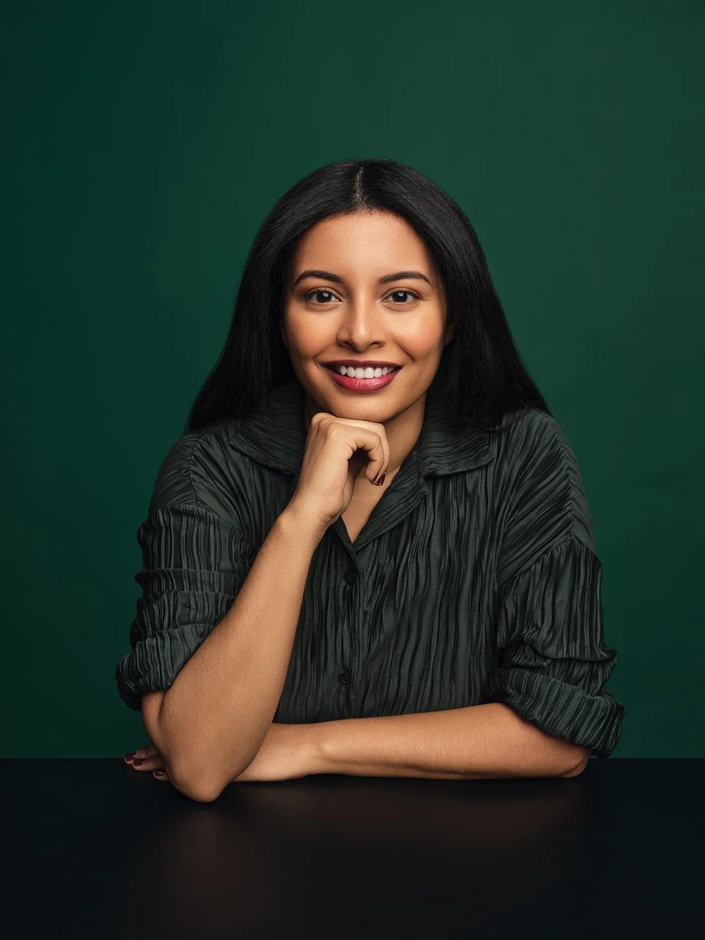 Headshot of a person with long black hair and a green shirt smiling with their chin resting on their hand against a dark green background.
