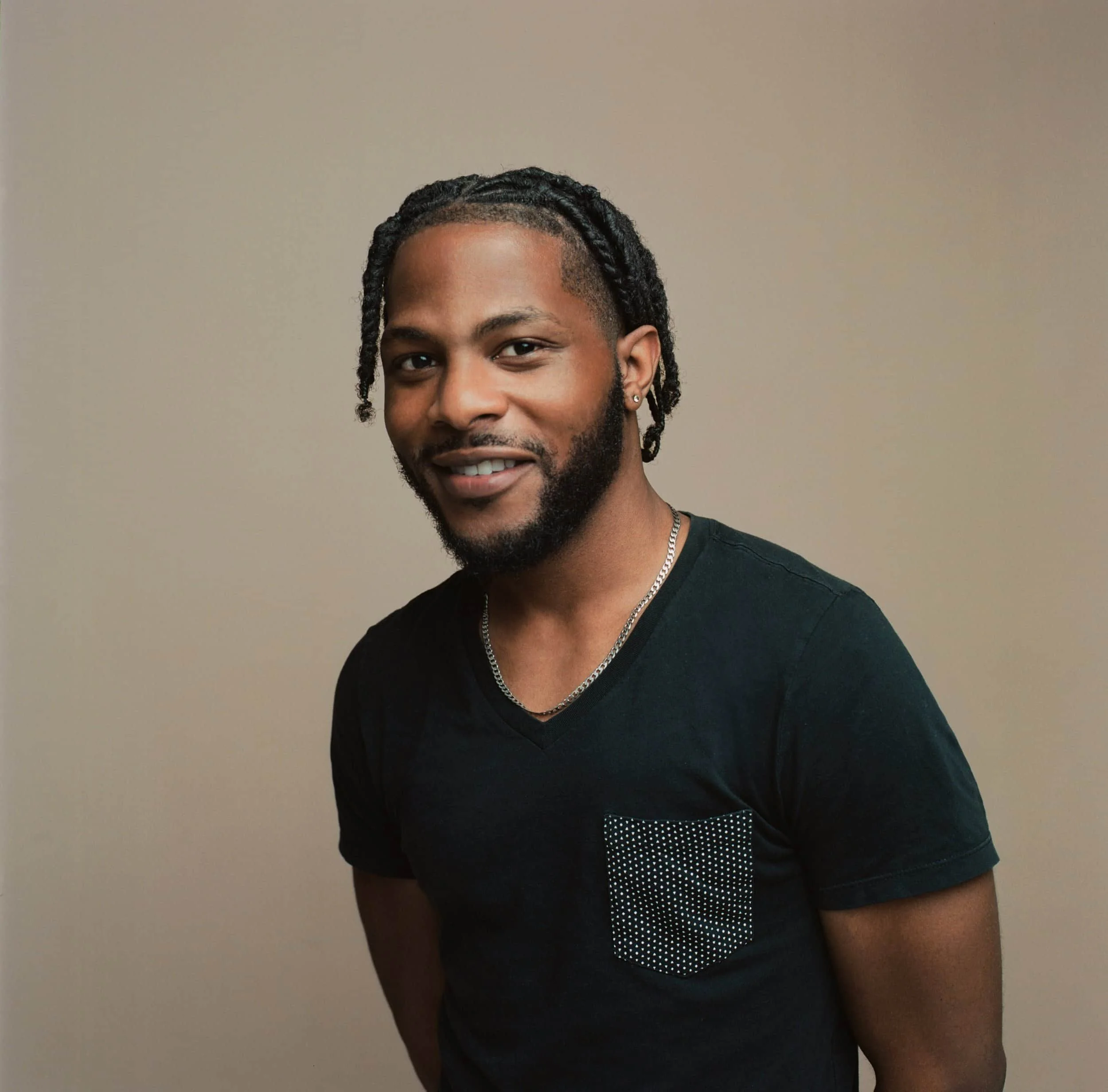 Young man with dark braided hair, beard, wearing earrings, black T-shirt, and silver necklace, standing against a plain beige background.