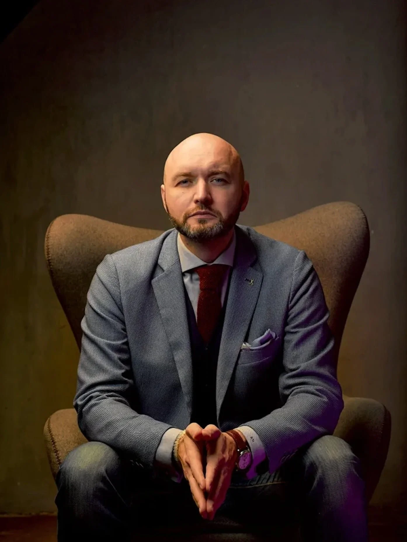 A bald man with a beard sitting on a brown upholstered armchair, wearing a light gray suit jacket, gray pants, a white shirt, and a dark red tie. He has a purple pocket square in his jacket pocket, a gold bracelet, a gold watch, and a wristband. His 