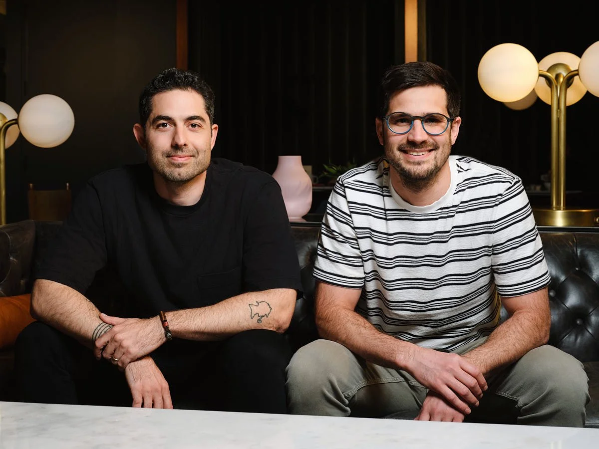 Two men are sitting on a black leather couch in a dimly lit room with modern decor. The man on the left has dark hair, a beard, and is wearing a black t-shirt. The man on the right has dark hair, glasses, a beard, and is wearing a striped t-shirt. Bo
