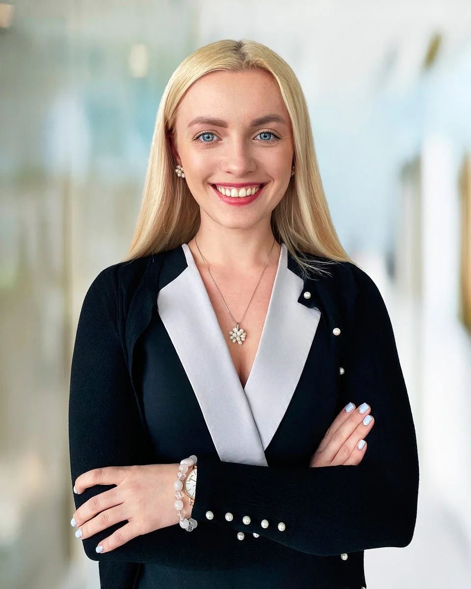 A young woman with long blonde hair, blue eyes, and a bright smile, wearing a black and white blazer with pearl accents, a flower-shaped necklace, pearl earrings, and a watch and bracelet, standing with her arms crossed in a well-lit corridor.