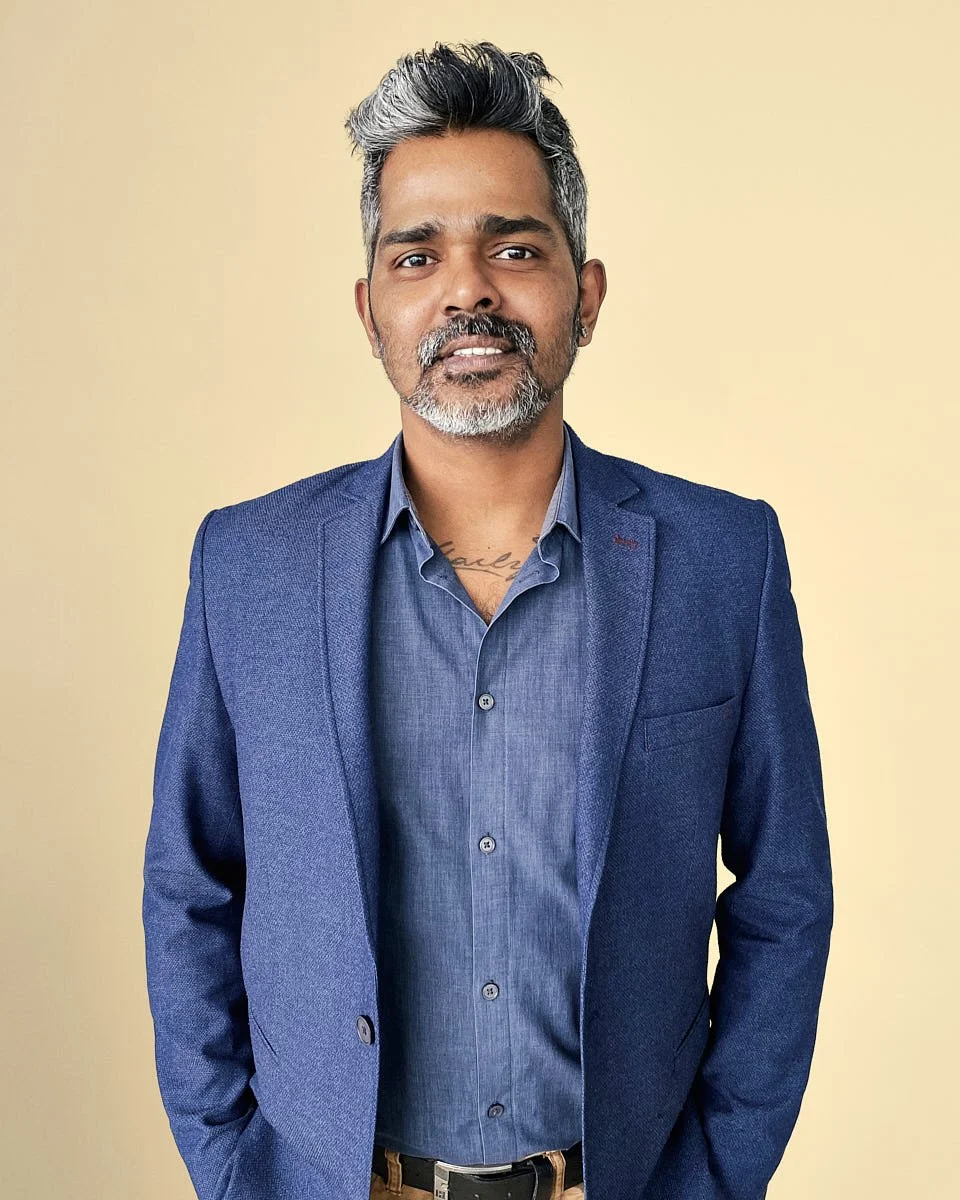 A man with gray hair and beard wearing a blue suit jacket and shirt, standing against a neutral beige background.