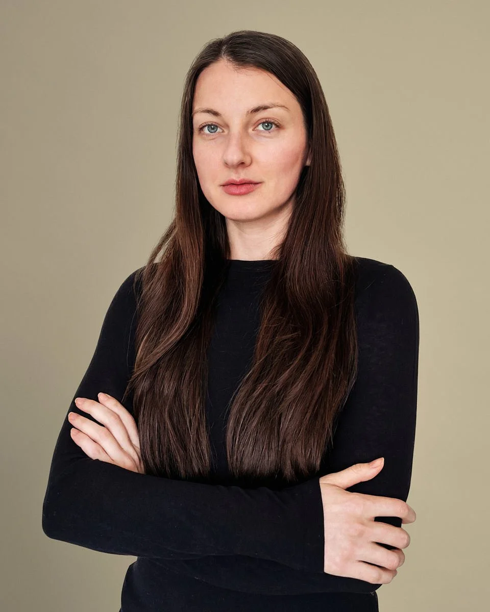 A woman with long brown hair and blue eyes wearing a black top standing against a beige background.