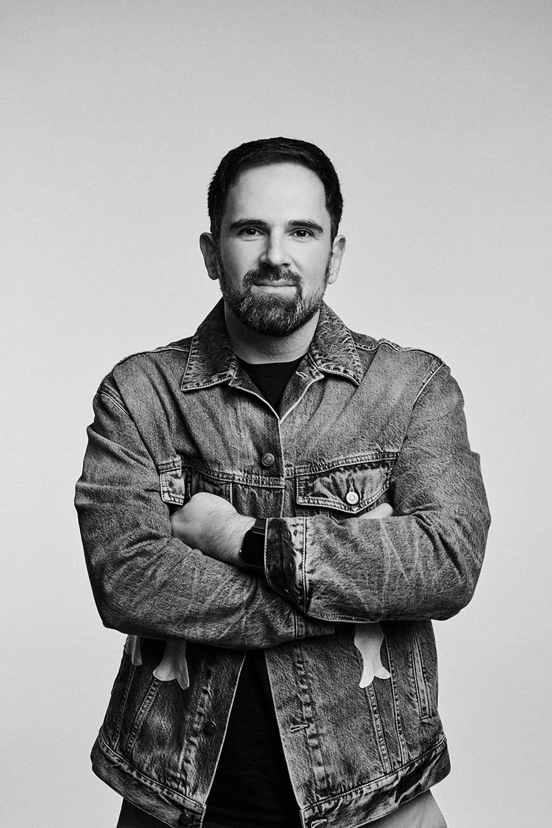 A black and white portrait of a man with dark hair and a beard, wearing a denim jacket and a black shirt, standing with arms crossed against a plain background.