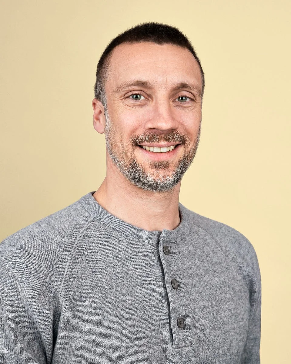 A smiling man with short brown hair, a beard, and light-colored eyes, wearing a gray henley shirt against a yellow background.