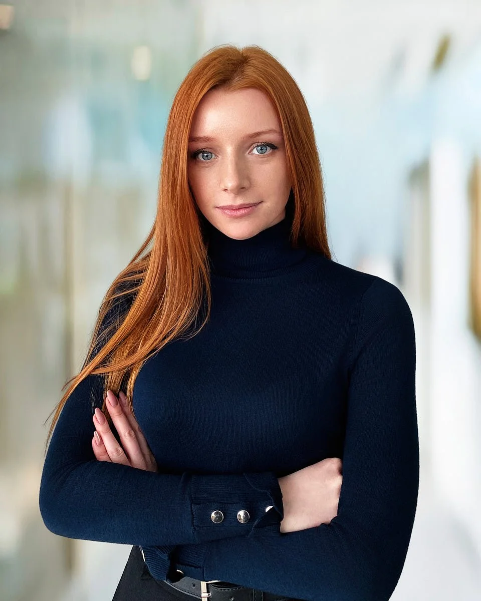A young woman with long red hair and blue eyes, wearing a navy turtleneck sweater, standing with arms crossed in an indoor setting with blurred background.