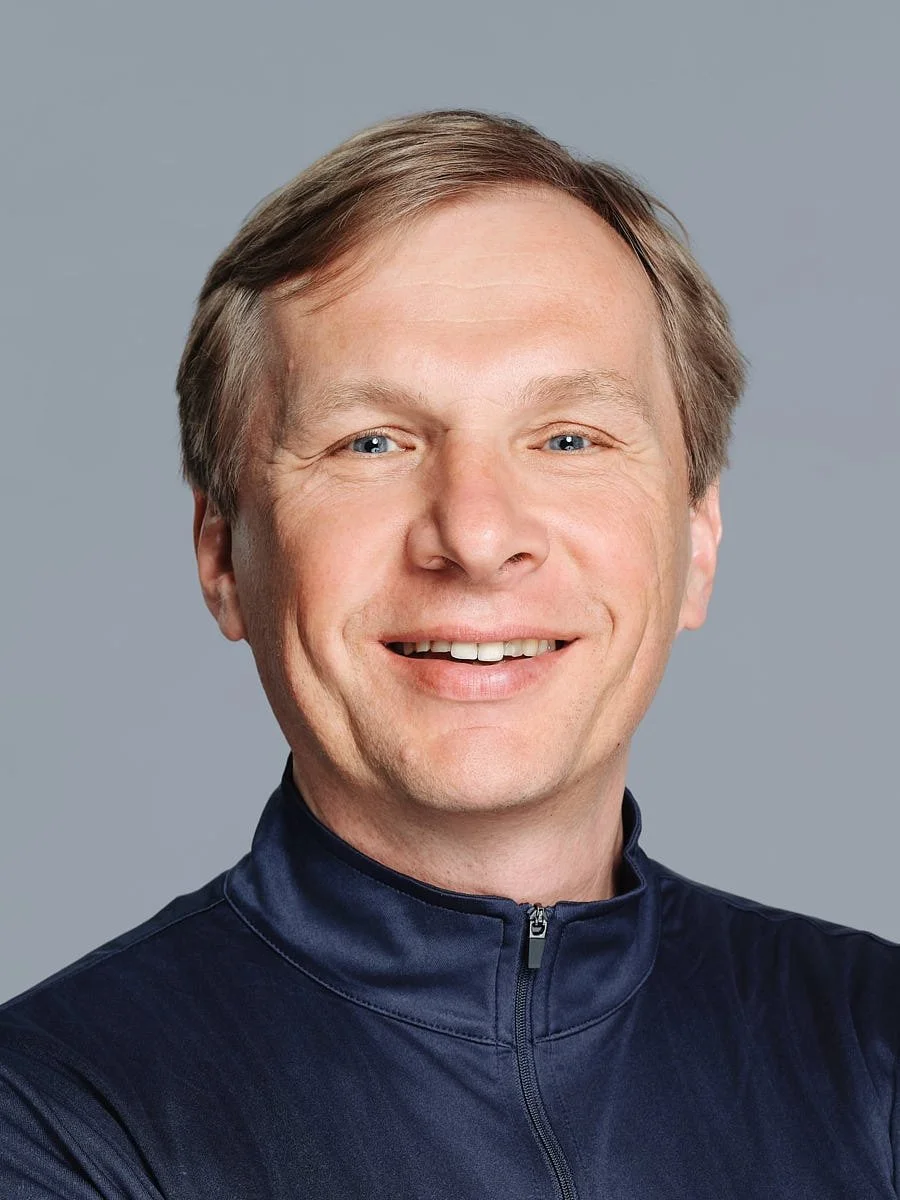 A smiling man with light skin, blonde hair, and blue eyes, wearing a navy blue zip-up athletic jacket, portrait against a plain gray background by Match Production NYC.