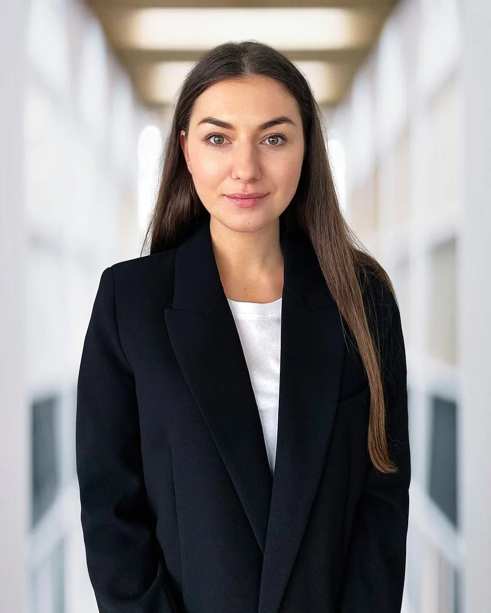 Professional woman with long brown hair wearing a black blazer and white shirt, standing in a bright corridor.