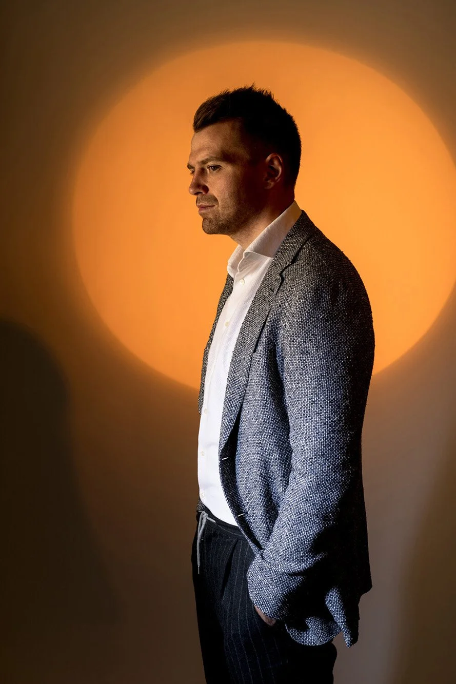 Man in textured blazer and white shirt standing in front of orange background.
