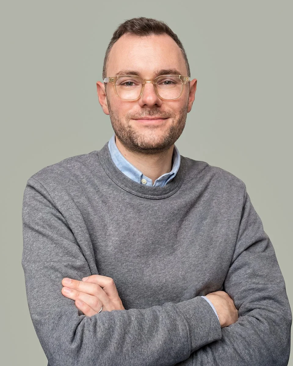 A man with short brown hair, glasses, and a beard wearing a gray sweater over a blue collared shirt, standing with arms crossed against a light green background.