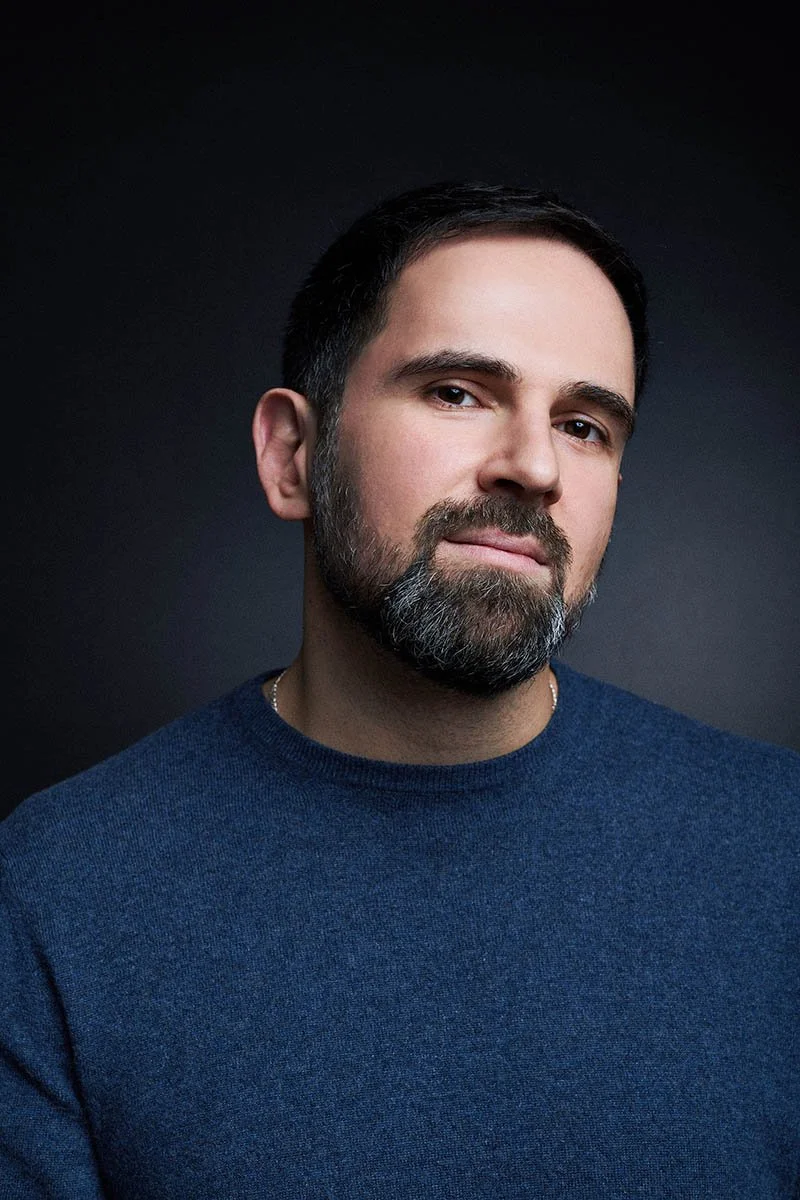 Portrait of a man with dark hair and beard, wearing a navy blue sweater, against a dark background.