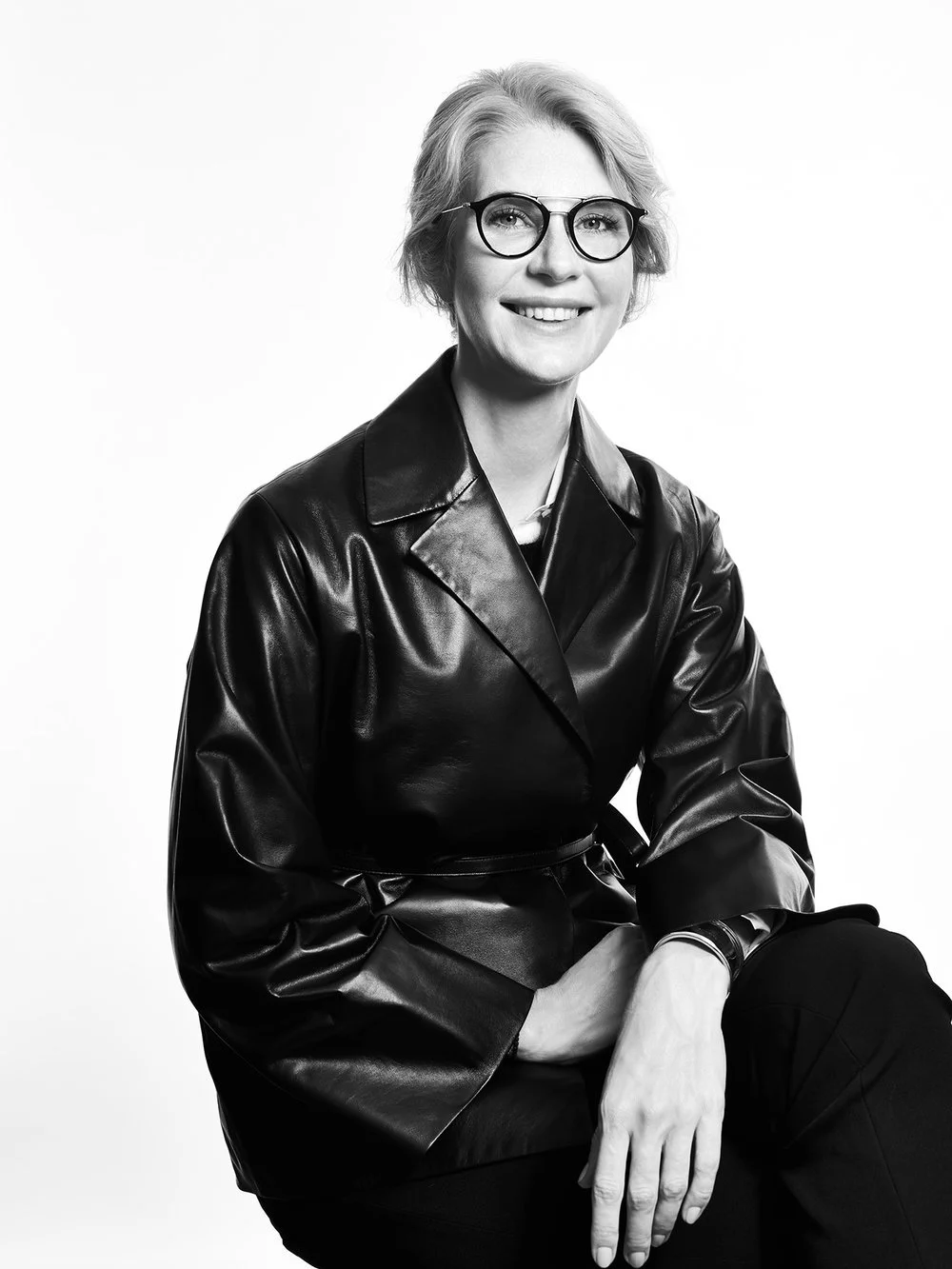 Black and white portrait of a woman with short hair, wearing glasses and a leather jacket, smiling and sitting with one arm resting on her knee.