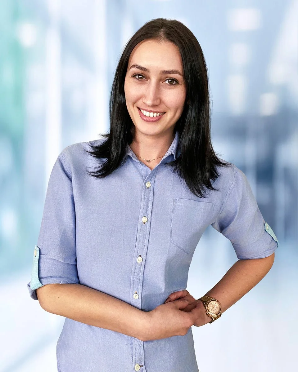 A young woman with black hair smiling, wearing a light blue button-up shirt with rolled-up sleeves, and a gold watch on her wrist, standing in front of a blurred background.