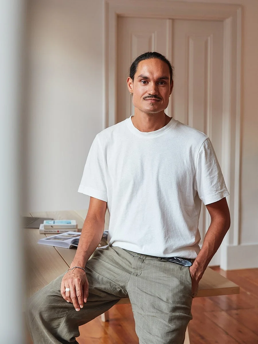 Man in a white T-shirt seated inside a warm-toned room.