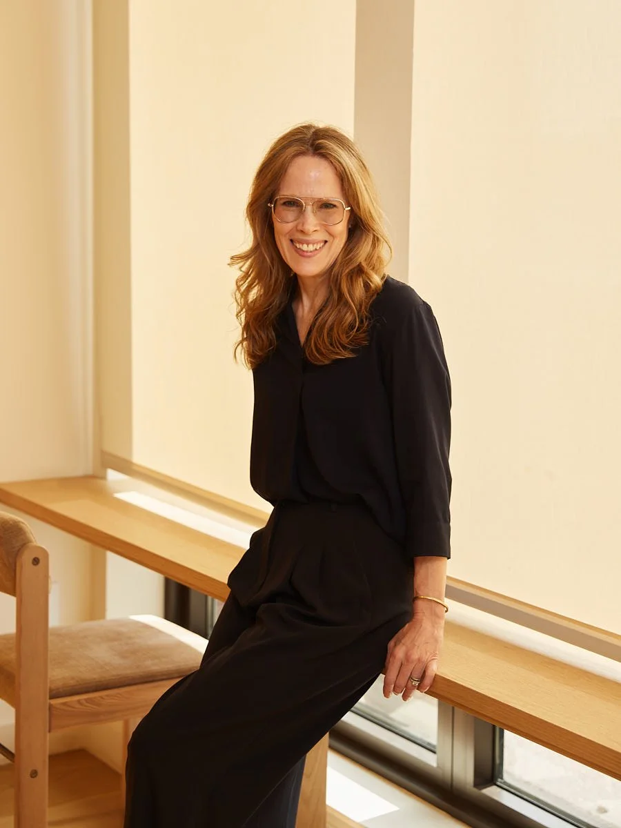 Woman in a dark outfit seated beside a sunlit window.