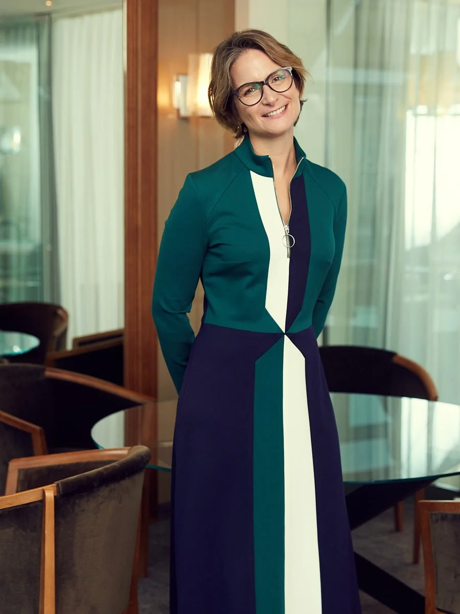 Woman wearing glasses and a teal dress standing in an office interior.