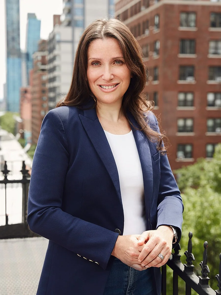 A woman in a navy blazer and white top stands beside a railing, with brick buildings behind her.