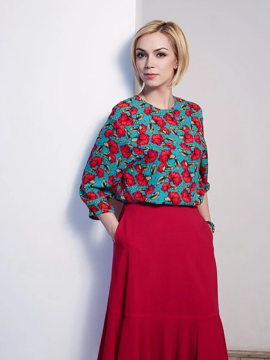 Woman in a colorful patterned top and red skirt against a pale background.