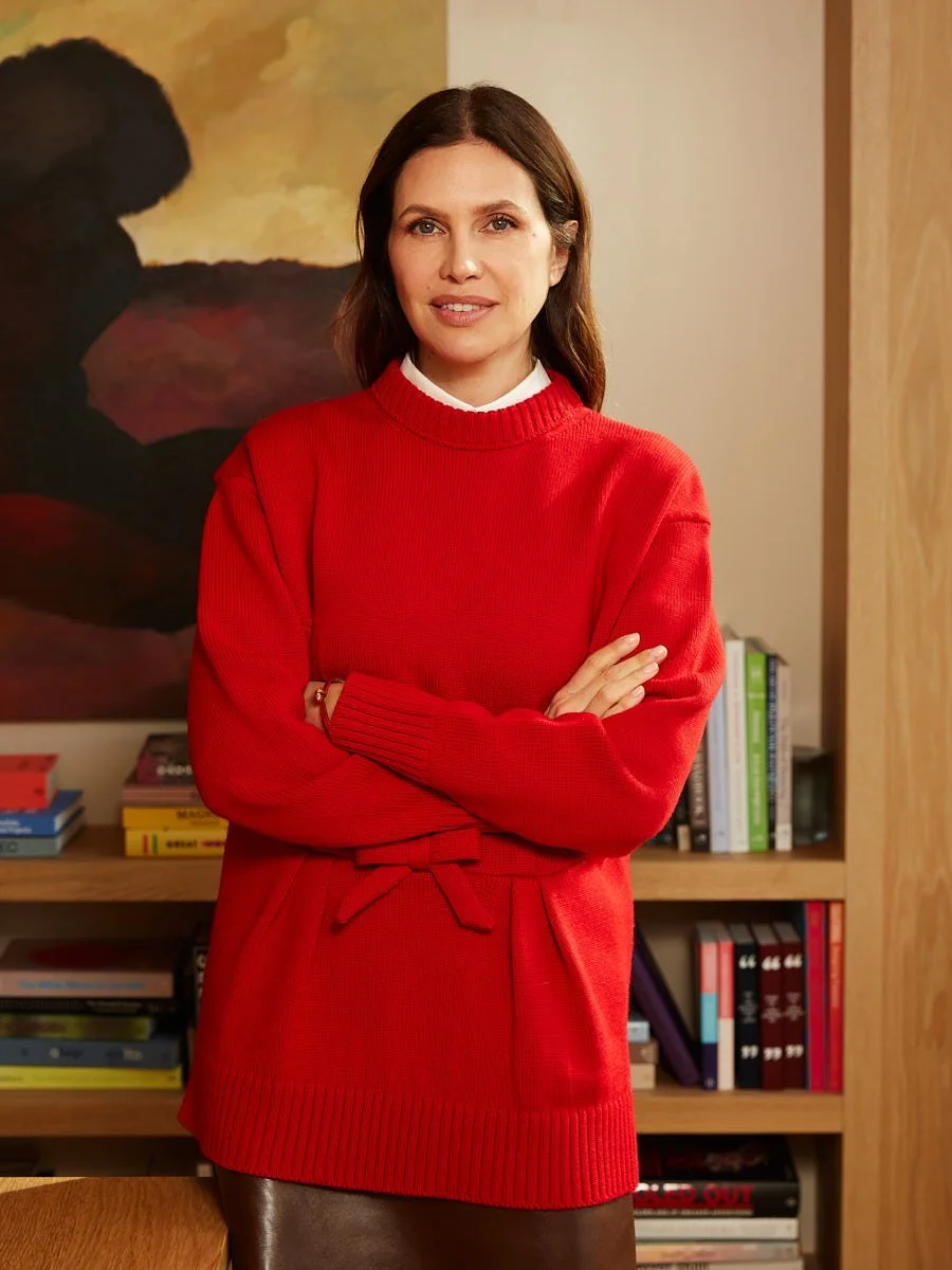 Woman in a red sweater standing with folded arms in front of bookshelves.