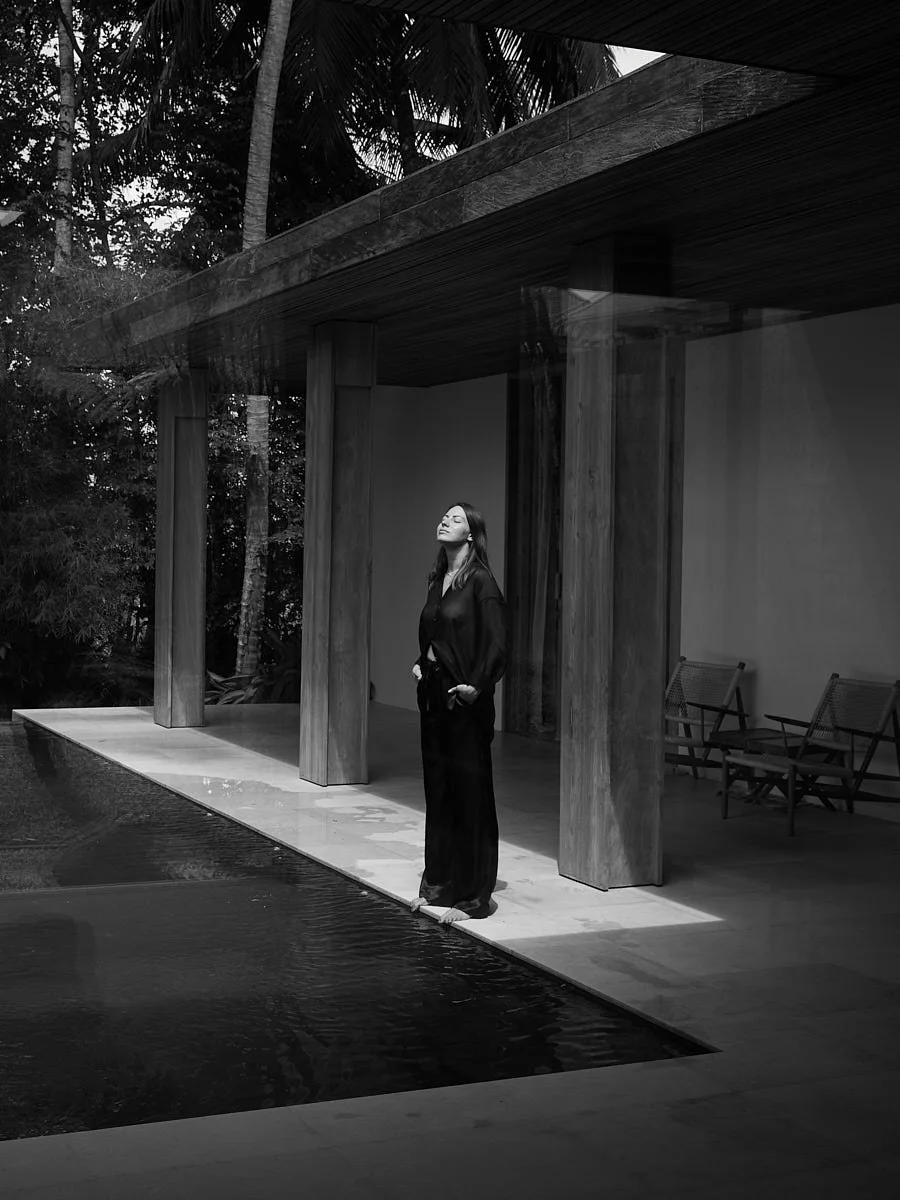 Woman in a dark outfit standing beside a pool beneath a modern building canopy.