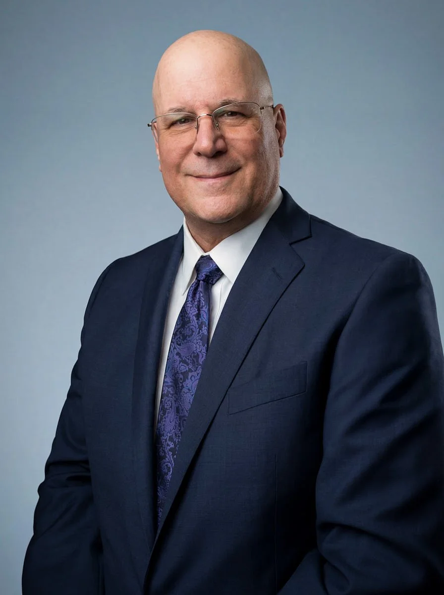 Bald man wearing glasses and a navy suit against a blue-gray background.