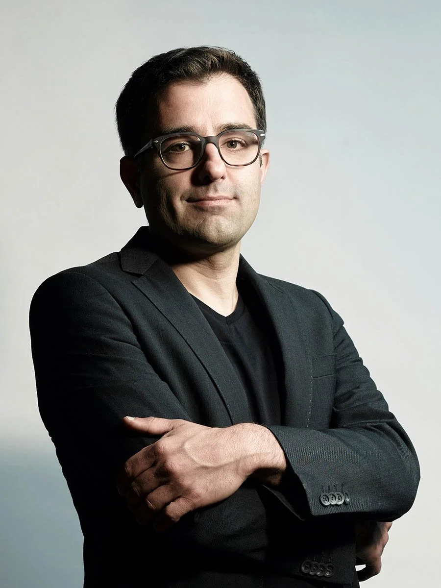 Portrait of a man with glasses in a dark blazer, standing with folded arms against a light background.