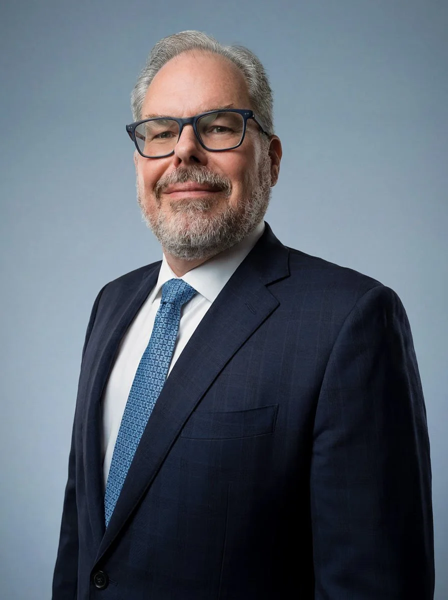 Man wearing glasses and a navy suit with a blue tie against a blue-gray background.