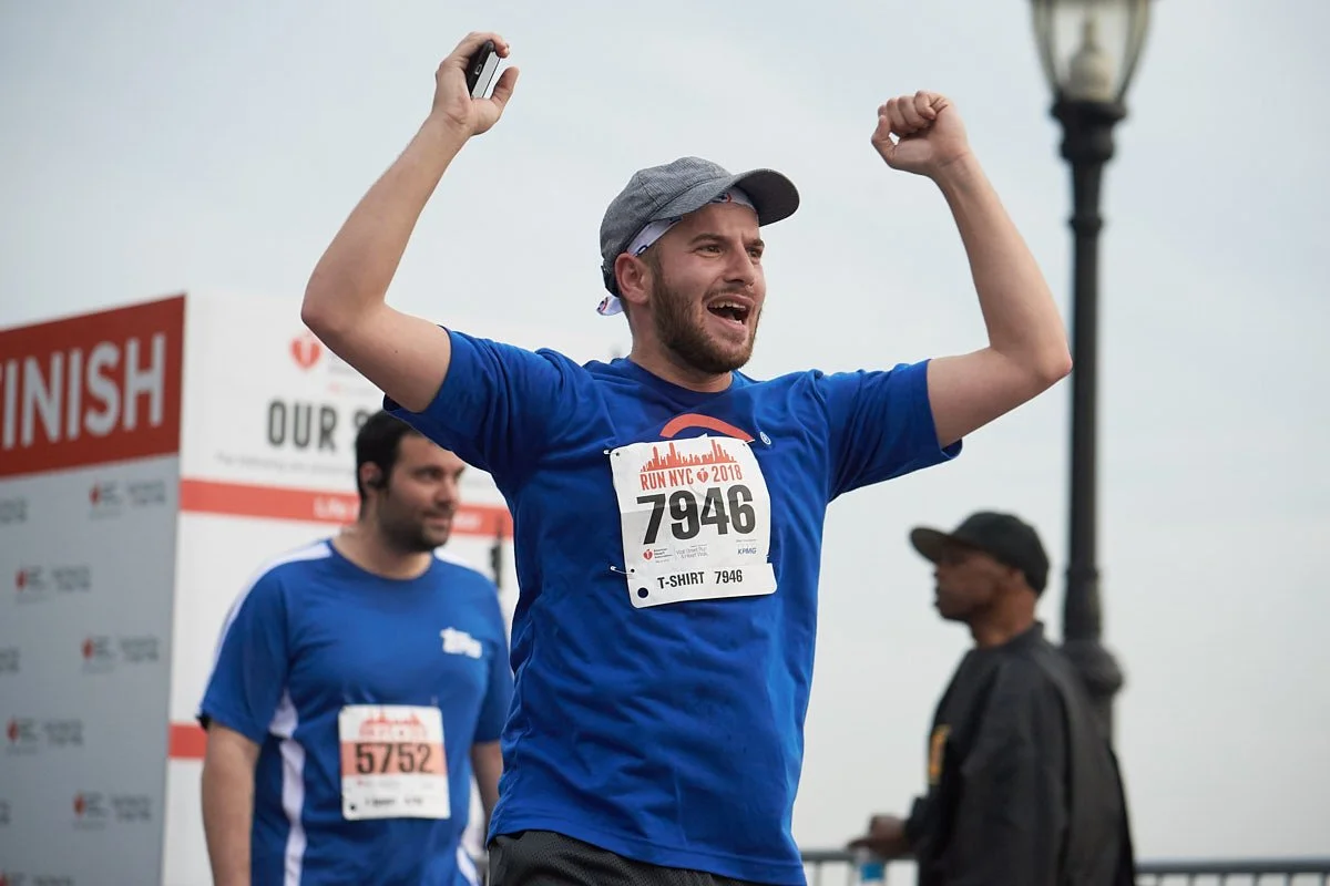 Runner in a blue shirt raises both arms beside a race finish banner.