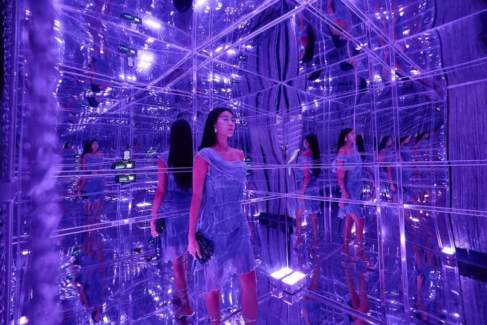 Woman standing among mirrored surfaces illuminated purple.