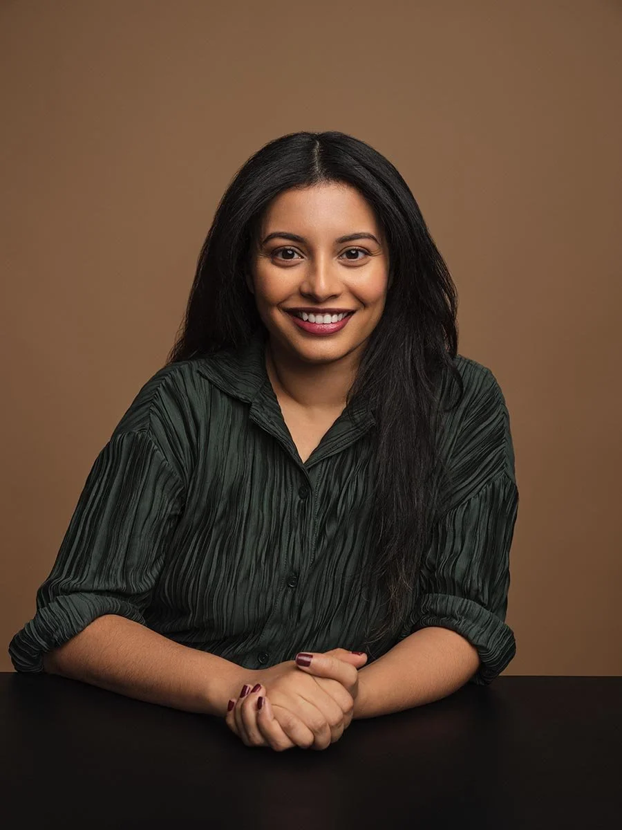 Smiling woman in a dark green shirt seated at a table against a brown background.