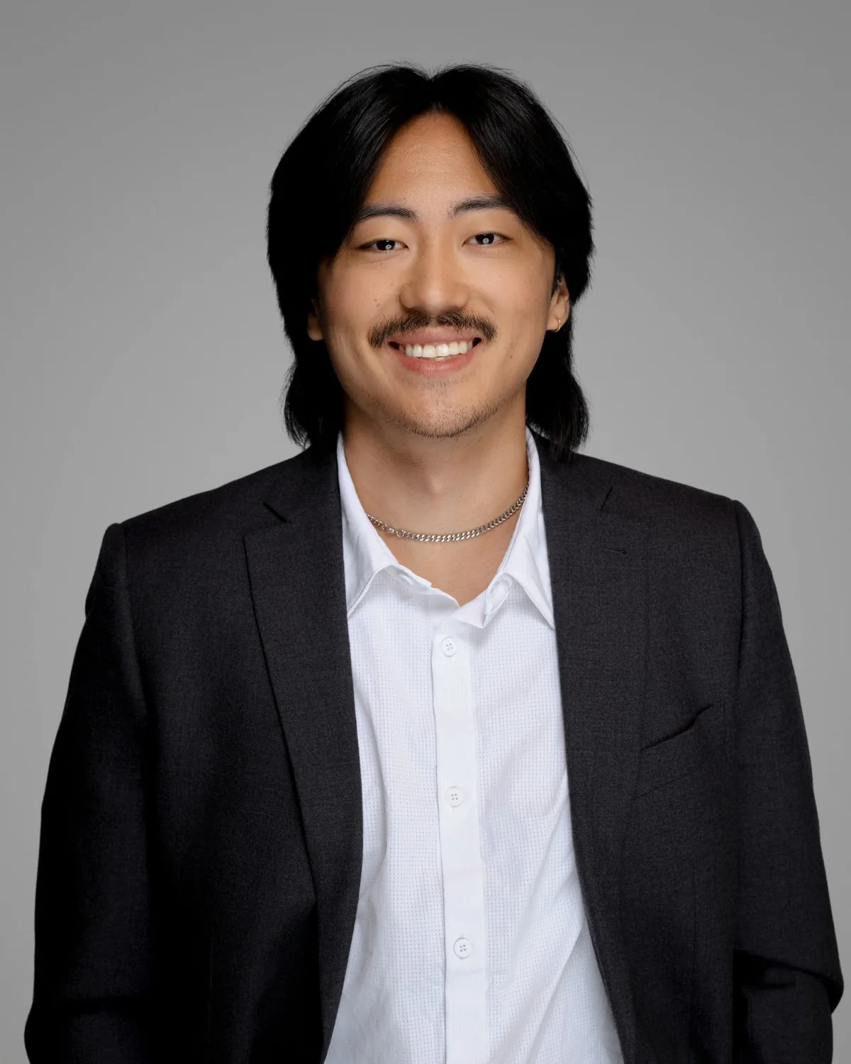 A person in a dark jacket and white shirt, smiling against a gray backdrop.