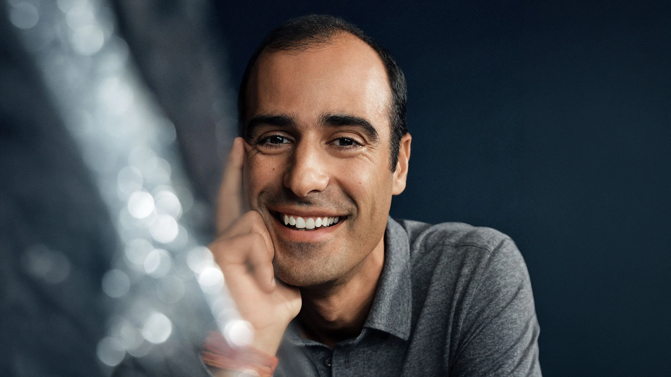 Man in a gray sweater smiling with one hand near his face against a dark background.